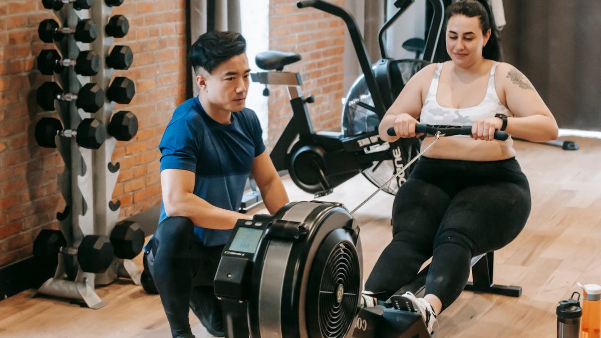 Positive young plus size female in leggings and crop top doing abs exercise on rowing machine during intense workout in modern gym with fit Asian male trainer